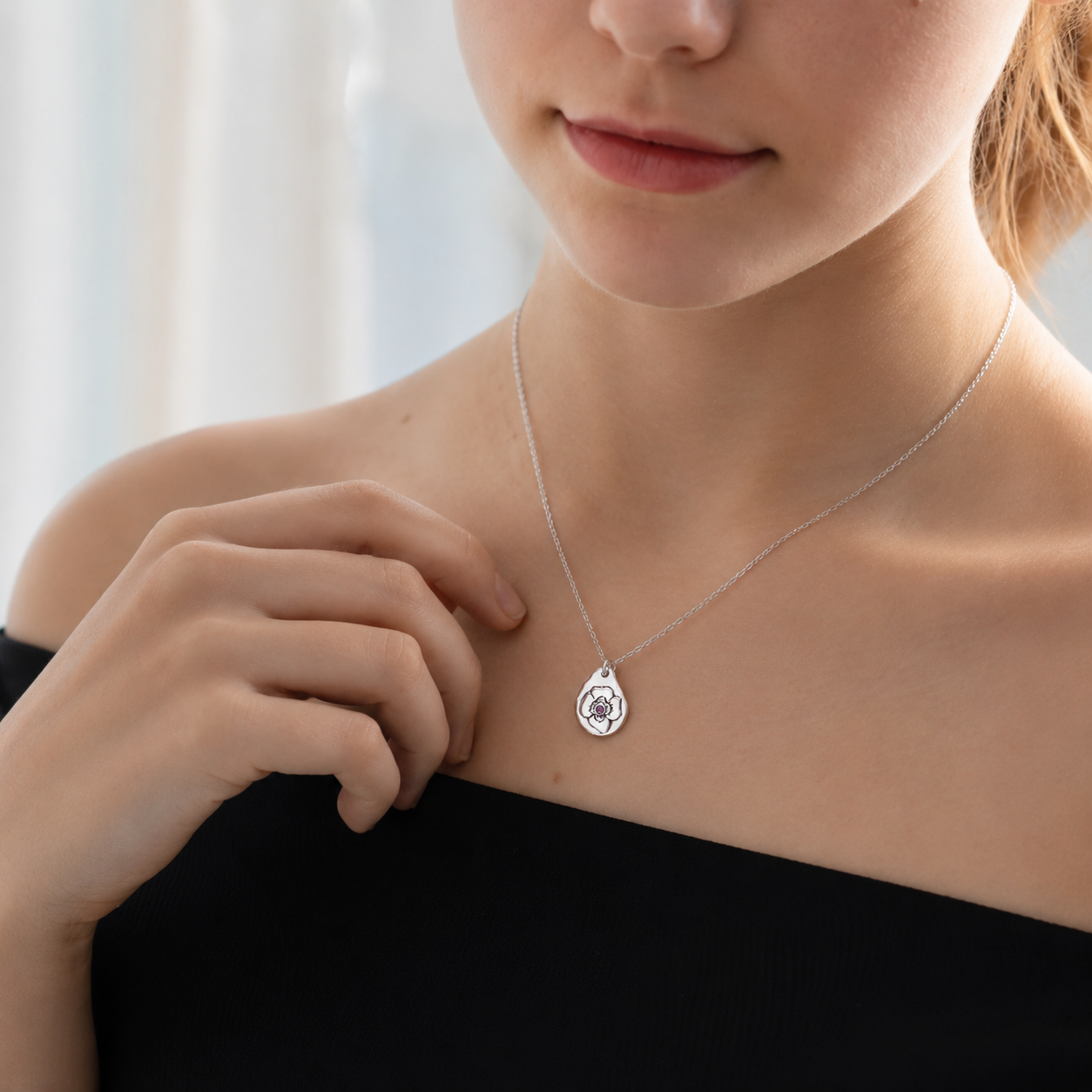 A close-up lifestyle shot of a model wearing a delicate silver teardrop pendant necklace with a circular emblem in the center, paired with a black off-the-shoulder top.