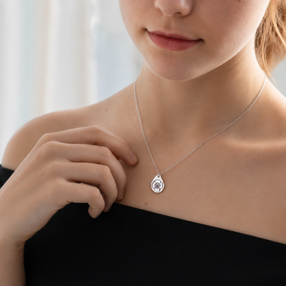 A close-up lifestyle shot of a model wearing a delicate silver teardrop pendant necklace with a circular emblem in the center, paired with a black off-the-shoulder top.