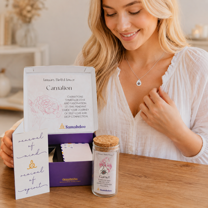 A woman in a white blouse sits at a wooden table with a Samahbdoo January Birth Flower set. The set includes a white card reading 'renunciation of mind', 'novelty of spirit', and 'carnalion', a purple box, and a small glass jar with a cork lid and white label.