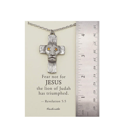 Silver cross necklace with lion head design on a card with text, next to a ruler for scale.