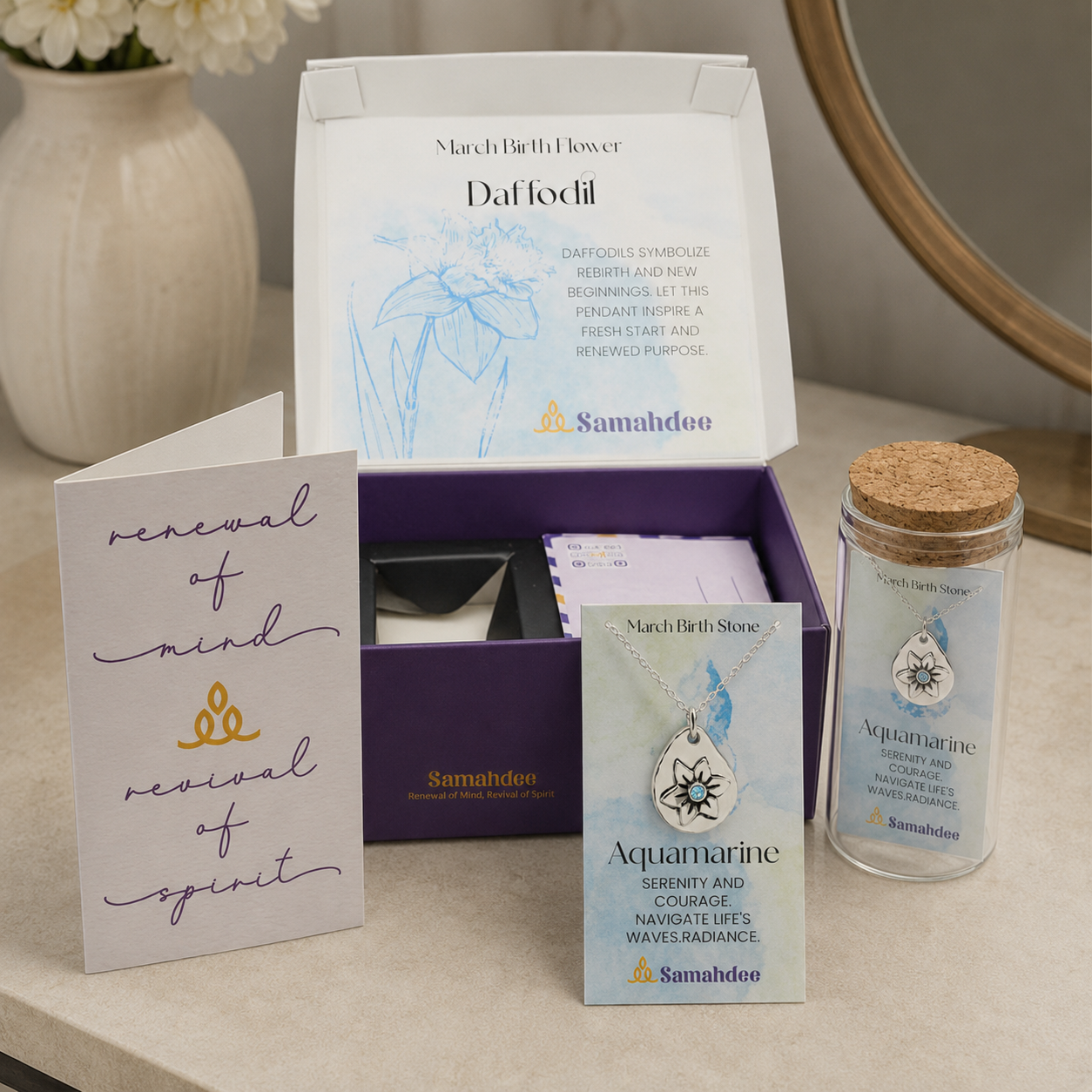 An open Samahdee gift box displaying a March Birth Flower card, a purple inner tray with folded cards, a small glass jar with a cork lid labeled Aquamarine, and a white card reading 'renewal of mind revival of spirit,' arranged on a light countertop with a vase and mirror in the background.