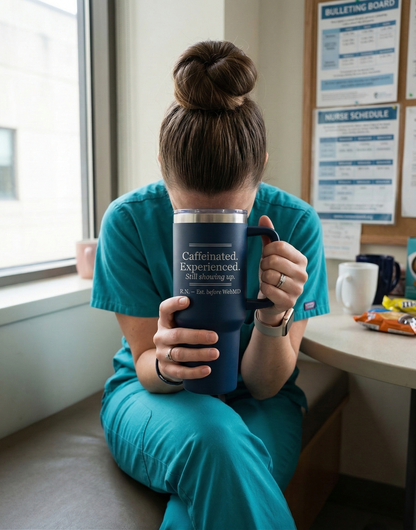 Person in teal scrubs holding a blue mug with text, sitting by a window.
