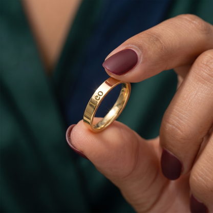 Gold ring held by a hand with maroon nail polish against a blurred green background
